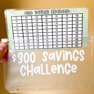May include: A clear plastic binder with a green and white chart inside. The chart has a floral pattern and the text "$300 Savings Challenge". The chart is divided into columns with dollar amounts: $1, $2, $5, and $10.