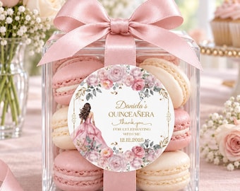 May include: A clear square box filled with pink and cream-colored macarons, tied with a dusty pink satin ribbon. A circular label features a floral wreath, a girl in a pink gown, and the text: Daniela's QUINCEAÑERA thank you FOR CELEBRATING WITH ME 12.12.2025.