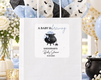 May include: A white paper gift bag with white handles, filled with tissue paper featuring black bats, spiders, and blue stars. A square label on the front displays a black cauldron with blue bubbles, text reading A BABY IS brewing, SAVANNAH'S Baby Shower, and 10.20.2025.