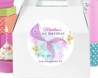 May include: White favor box with a round sticker featuring a mermaid tail with purple, blue, and gold glitter accents. The sticker says "Madeline's 1st Birthday" and "Thank You for Splashing By".