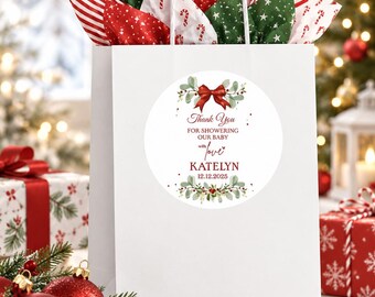 May include: A white gift bag with white twisted paper handles, filled with red, green, and white patterned tissue paper. A circular sticker on the front features a red bow, greenery, and the text Thank You FOR SHOWERING OUR BABY with love KATELYN 12.12.2025.