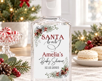 May include: A clear plastic hand sanitizer bottle with a white flip-top cap. The label features a festive botanical design with pine branches, red berries, and pinecones. Text reads: SANTA baby, Amelia's Baby Shower, and the date 12.12.2025.