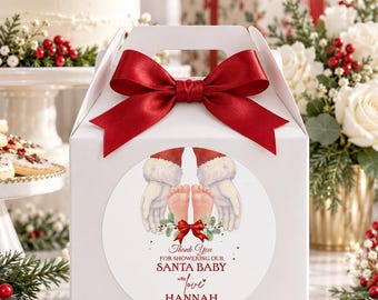 May include: A white gable box featuring a large red satin bow and a circular sticker. The sticker displays Santa's gloved hands cradling baby feet, with text reading: Thank You FOR SHOWERING OUR SANTA BABY with love HANNAH 12.20.2025.