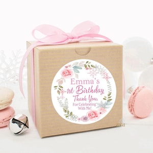 May include: A small brown gift box with a pink ribbon and a round sticker. The sticker has a white background with a floral wreath and snowflakes. The text on the sticker reads "Emma's 1st Birthday Thank You. For Celebrating With Me!"