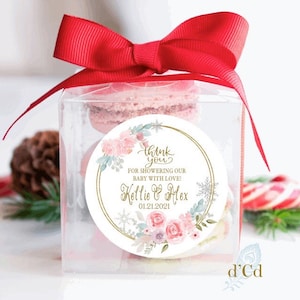 May include: A clear box with a red bow tied on top. Inside the box are pink macarons. A round sticker with a gold border and floral design says "Thank you for showering our baby with love! Kellie & Alex 01.21.2021"