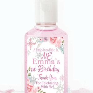 May include: A pink hand sanitizer bottle with a white cap. The label reads "A Little Snowflake Is, NE Emma's 1st Birthday Thank You, For Celebrating With Me!" with floral and snowflake accents. A birthday party favor.