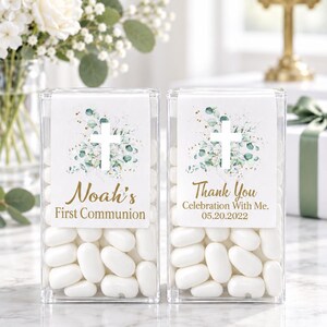 May include: Two clear rectangular plastic containers filled with white oval candies. Each features a white label with a green eucalyptus leaf design, a white cross, and gold text. One reads Noah's First Communion, the other Thank You Celebration With Me 05.20.2022.