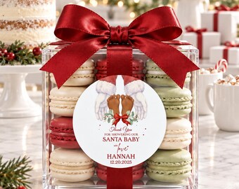 May include: A clear square gift box filled with red, white, and light green macarons, topped with a large dark red satin ribbon bow. A circular white label features an illustration of Santa hands holding baby feet with the text Thank You FOR SHOWERING OUR SANTA BABY with love HANNAH 12.20.2025.