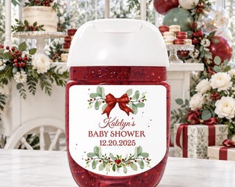 May include: A clear plastic hand sanitizer bottle filled with red gel, featuring a white label with a red bow, green foliage, and the text Katelyn's BABY SHOWER 12.20.2025. The bottle has a white flip-top cap and a rounded rectangular shape.