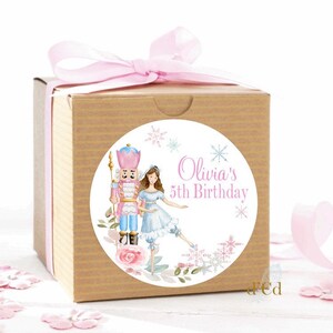 May include: A round, white sticker with a watercolor illustration of a Nutcracker and a ballerina. The sticker has pink and blue accents and says "Olivia's 5th Birthday" in pink text.