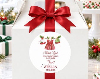 May include: A white cardboard gift box with a handle, tied with a red satin ribbon bow. A circular sticker on the front features a watercolor illustration of a red dress and two hats hanging on a clothesline, with the text Thank You FOR SHOWERING OUR BABY with love STELLA 12.12.2025.