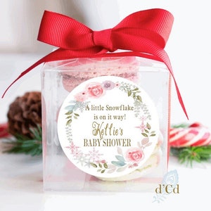 May include: A white round favor tag with a floral wreath design and gold text that reads "A little Snowflake is on it's way! Keltie's Baby Shower". The tag is attached to a clear box with a red ribbon and a stack of macarons inside.
