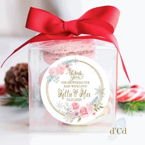 May include: A clear box with a red bow tied on top. Inside the box are pink macarons. A round sticker with a gold border and floral design says "Thank you for showering our baby with love! Kellie & Alex 01.21.2021"