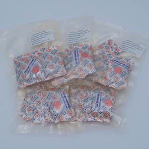 Puede incluir: Primer plano de una pila de pequeñas bolsas blancas selladas con un círculo rojo en el centro. Las bolsas están etiquetadas como "Absorbedor de oxígeno" y tienen un certificado de calidad con un número de lote.