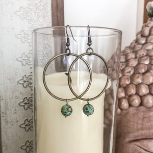 Puede incluir: Un par de pendientes de aro con cuentas de color turquesa que cuelgan de la parte inferior. Los aros son de metal de color latón.