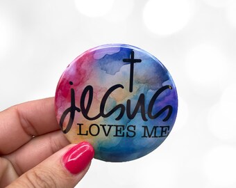 May include: A round button with a watercolor background of pink, blue, and purple hues. The button features the words "Jesus Loves Me" in black script, with a black cross above the word "Jesus".