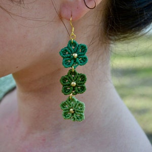 May include: Green and gold flower-shaped earrings with three flowers on each earring. The flowers are made of macrame and have gold beads in the center.