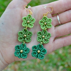 Pode incluir: Um par de brincos de flores verdes e douradas com três flores em cada brinco. As flores são feitas de macramé e têm uma conta dourada no centro.