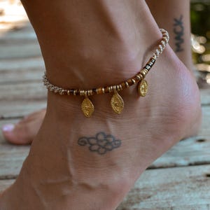 May include: A white and brown beaded ankle bracelet with gold leaf charms. The bracelet is worn on a person's ankle.