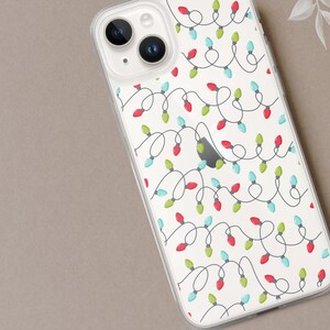 May include: Clear phone case with a repeating pattern of Christmas lights. The lights are red, green, and blue, connected by a dark gray wire, set against a white background. The phone case is on a neutral-toned surface.
