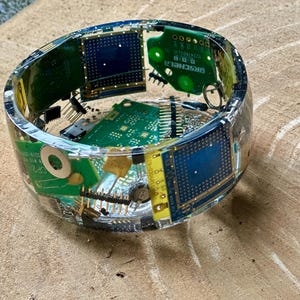 May include: A clear resin bangle bracelet embedded with colorful electronic circuit board components. The bracelet features green, blue, and yellow circuit boards, with various chips and connectors visible. The bangle is approximately 3 inches in diameter.