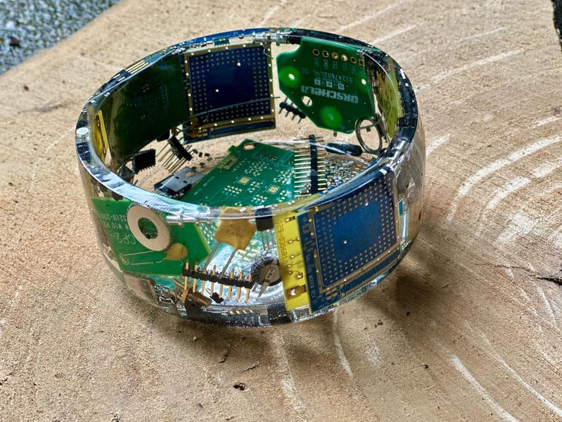 K&ouml;nnte beinhalten: Ein Armreif aus klarem Harz, in den farbenfrohe elektronische Leiterplattenkomponenten eingebettet sind. Der Armreif weist gr&uuml;ne, blaue und gelbe Leiterplatten auf, mit sichtbaren Chips und Anschl&uuml;ssen. Der Armreif hat einen Durchmesser von etwa 7,6 cm.