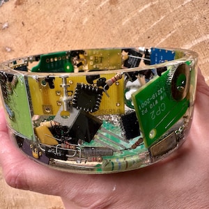 K&ouml;nnte beinhalten: Eine transparente Harzschale gef&uuml;llt mit bunten elektronischen Bauteilen, darunter Leiterplatten, Widerst&auml;nde und Chips. Die Schale wird in einer Hand gehalten und zeigt die eingebettete Technologie und ihr einzigartiges Design.