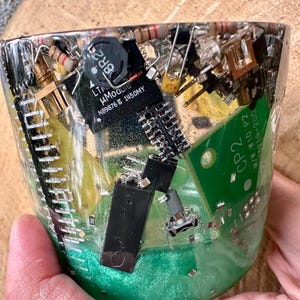 K&ouml;nnte beinhalten: Eine transparente Harz-Tasse gef&uuml;llt mit verschiedenen elektronischen Bauteilen, darunter Leiterplatten, Widerst&auml;nde und Mikrochips. Der Boden der Tasse hat einen gr&uuml;nen Farbton. Die Tasse wird von einer Person gehalten.
