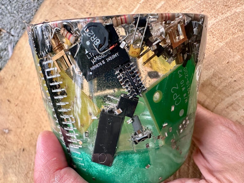 K&ouml;nnte beinhalten: Eine transparente Harz-Tasse gef&uuml;llt mit verschiedenen elektronischen Bauteilen, darunter Leiterplatten, Widerst&auml;nde und Mikrochips. Der Boden der Tasse hat einen gr&uuml;nen Farbton. Die Tasse wird von einer Person gehalten.