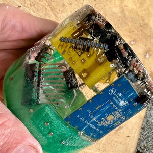 K&ouml;nnte beinhalten: Ein klarer Harzblock umschlie&szlig;t verschiedene elektronische Komponenten, darunter gelbe und blaue Leiterplatten, Widerst&auml;nde und andere Kleinteile. Der Block hat eine gr&uuml;ne Basis und wird von einer Hand gehalten.