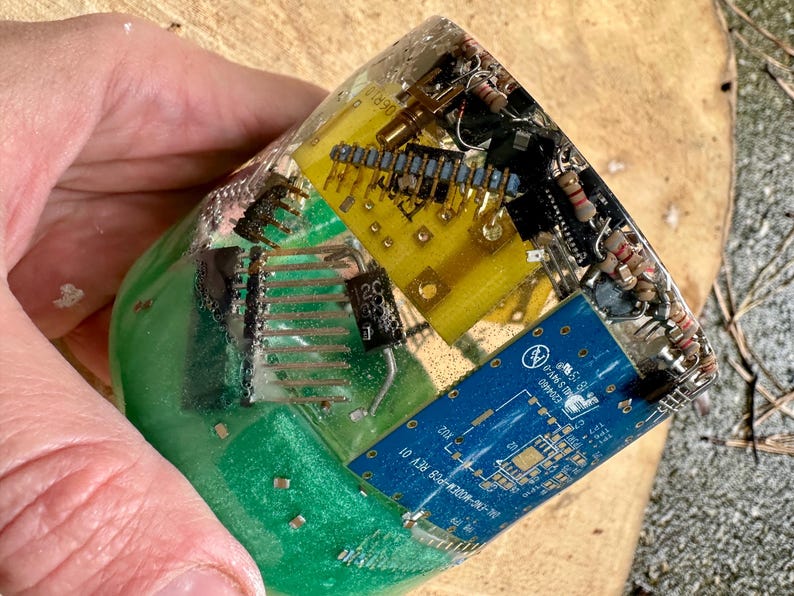 K&ouml;nnte beinhalten: Ein klarer Harzblock umschlie&szlig;t verschiedene elektronische Komponenten, darunter gelbe und blaue Leiterplatten, Widerst&auml;nde und andere Kleinteile. Der Block hat eine gr&uuml;ne Basis und wird von einer Hand gehalten.