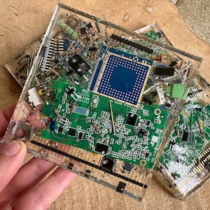 May include: Clear resin coasters embedded with electronic components, including green circuit boards, chips, and various small parts. The coasters have a square shape and are displayed on a wooden surface.
