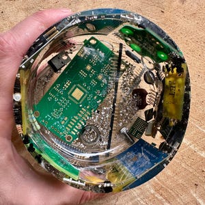 K&ouml;nnte beinhalten: Ein klarer, runder Aschenbecher aus Kunstharz mit eingebetteten elektronischen Bauteilen, darunter gr&uuml;ne Leiterplatten und silberne Metallteile. Der Aschenbecher wird in einer Hand gehalten.