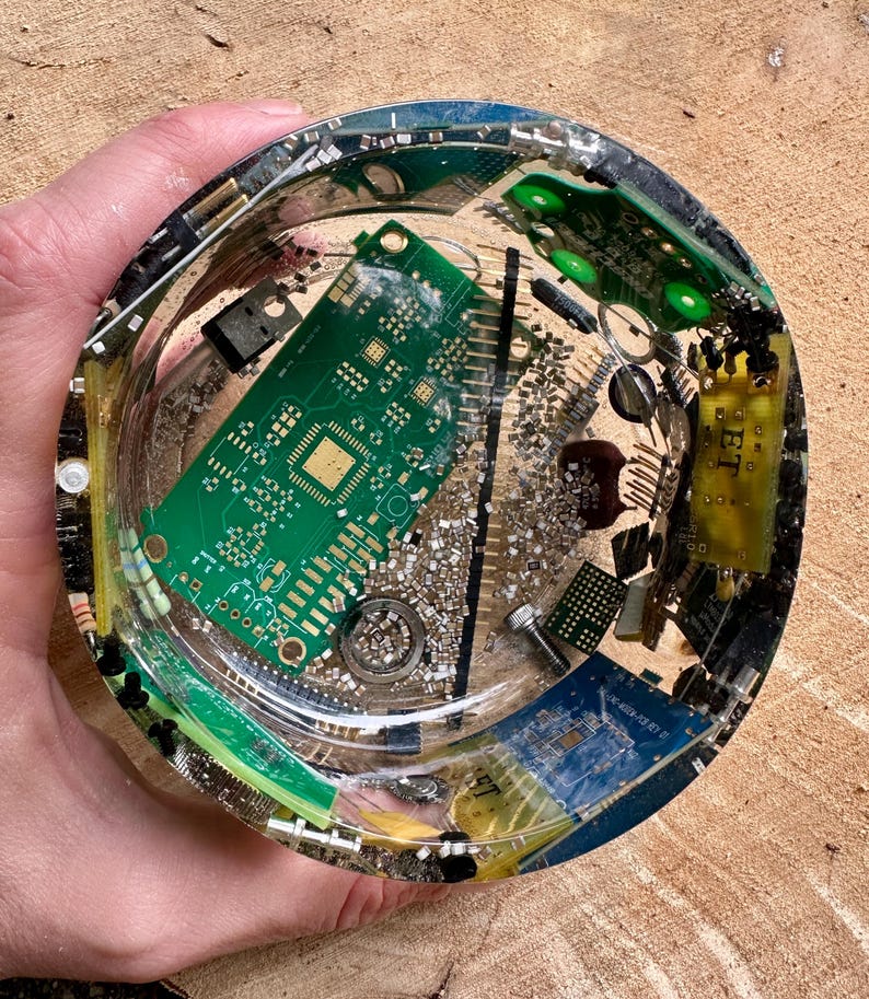 K&ouml;nnte beinhalten: Ein klarer, runder Aschenbecher aus Kunstharz mit eingebetteten elektronischen Bauteilen, darunter gr&uuml;ne Leiterplatten und silberne Metallteile. Der Aschenbecher wird in einer Hand gehalten.