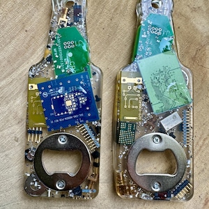 May include: Two bottle-shaped bottle openers made of clear resin embedded with various electronic components, including green and blue circuit boards, resistors, and metal pins. Each opener features a circular metal insert at the base.