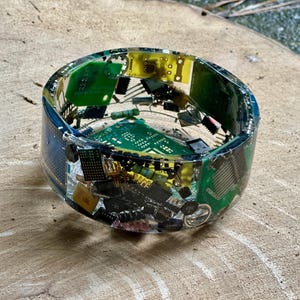 K&ouml;nnte beinhalten: Ein Armreif aus klarem Harz, in den farbenfrohe elektronische Bauteile eingebettet sind, darunter gr&uuml;ne Leiterplatten, gelbe Chips und verschiedene kleine schwarze und silberne Teile. Das Armband hat eine runde Form und wird auf einer Holzoberfl&auml;che pr&auml;sentiert.