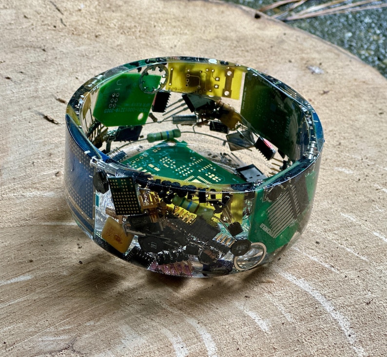 K&ouml;nnte beinhalten: Ein Armreif aus klarem Harz, in den farbenfrohe elektronische Bauteile eingebettet sind, darunter gr&uuml;ne Leiterplatten, gelbe Chips und verschiedene kleine schwarze und silberne Teile. Das Armband hat eine runde Form und wird auf einer Holzoberfl&auml;che pr&auml;sentiert.