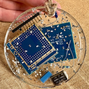 May include: A clear, circular ornament with embedded blue circuit boards, electronic components, and silver flecks. A brown twine loop is attached for hanging. The ornament features a variety of electronic parts.