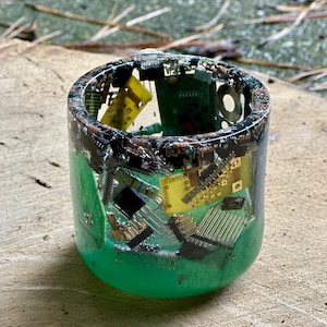 K&ouml;nnte beinhalten: Ein zylindrisches Kunstwerk aus Harz mit gr&uuml;nem Sockel, das verschiedene elektronische Bauteile umschlie&szlig;t. Die Oberseite ist mit einer Mischung aus Leiterplatten, Chips und anderen Kleinteilen gef&uuml;llt. Das St&uuml;ck steht auf einer Holzoberfl&auml;che.