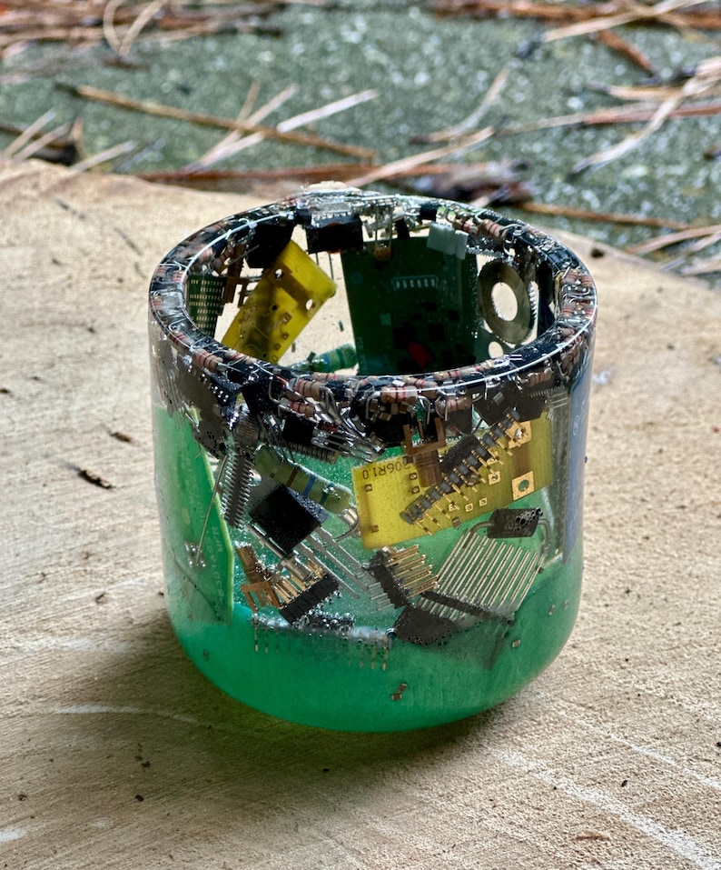 K&ouml;nnte beinhalten: Ein zylindrisches Kunstwerk aus Harz mit gr&uuml;nem Sockel, das verschiedene elektronische Bauteile umschlie&szlig;t. Die Oberseite ist mit einer Mischung aus Leiterplatten, Chips und anderen Kleinteilen gef&uuml;llt. Das St&uuml;ck steht auf einer Holzoberfl&auml;che.
