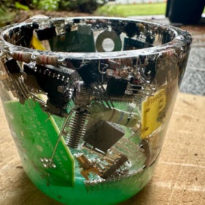 K&ouml;nnte beinhalten: Ein klarer Becher aus Kunstharz, der verschiedene elektronische Bauteile enth&auml;lt, darunter gr&uuml;ne Leiterplatten, Widerst&auml;nde und schwarze Chips. Der Becher hat eine abgerundete Form und eine glatte, gl&auml;nzende Oberfl&auml;che.