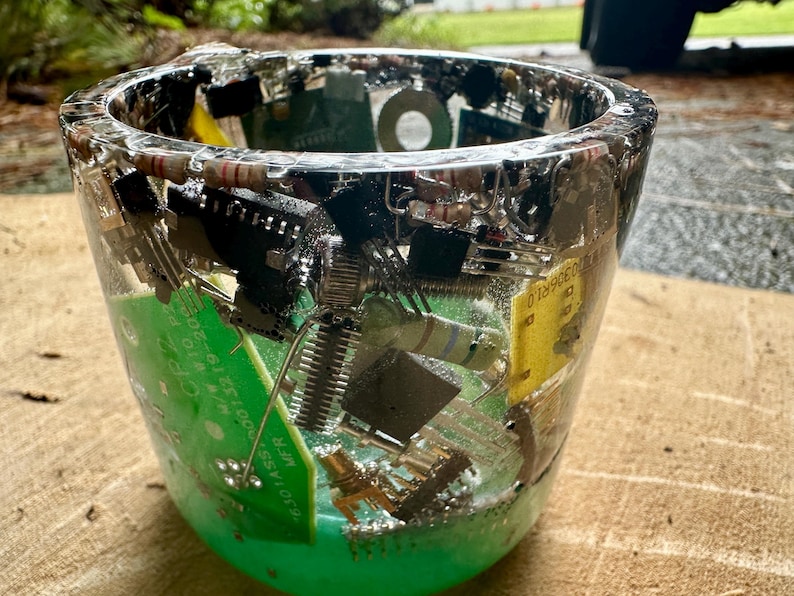 K&ouml;nnte beinhalten: Ein klarer Becher aus Kunstharz, der verschiedene elektronische Bauteile enth&auml;lt, darunter gr&uuml;ne Leiterplatten, Widerst&auml;nde und schwarze Chips. Der Becher hat eine abgerundete Form und eine glatte, gl&auml;nzende Oberfl&auml;che.