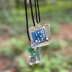 May include: A necklace featuring two clear resin pendants containing embedded electronic circuit board components. The larger pendant is square, and the smaller one is rectangular. Both are suspended from a black braided cord necklace.