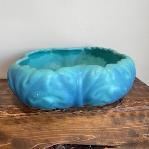 May include: A turquoise ceramic bowl with a textured, organic shape. The bowl has a wavy rim and a gradient of blue hues, from a lighter shade at the top to a deeper tone at the base. The bowl is displayed on a wooden surface.