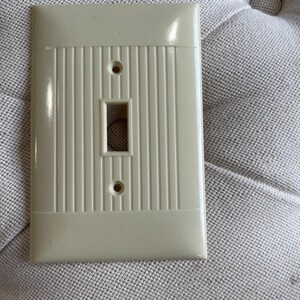 May include: Off-white, vintage-style light switch cover. The cover has a rectangular shape with a vertical ribbed design in the center and a rectangular opening for the switch. Two small holes are located above and below the switch opening.