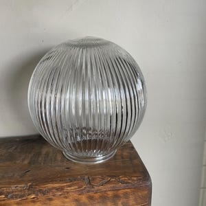 May include: A clear, ribbed glass globe, likely a light fixture shade, sits on a wooden surface. The globe has a spherical shape with vertical ridges, creating a textured appearance. The glass is transparent, allowing light to pass through.