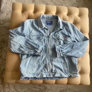 May include: Light blue denim jacket with a zip-up front, buttoned cuffs, and chest pockets. The jacket has a collar and a label that reads "Pendleton". The jacket is laid on a beige tufted ottoman.