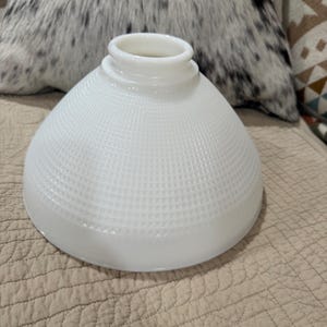 May include: A white milk glass lamp shade with a conical shape and a textured grid pattern across its surface. The shade features a circular opening at the top with a raised rim designed for mounting, and a wide, smooth edge at the base.