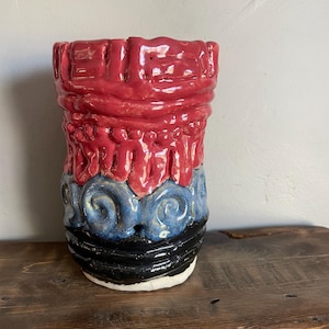 May include: A cylindrical ceramic vase featuring a glossy finish with three distinct horizontal bands of color. The top section is bright red with raised linear textures, the middle is blue with swirling patterns, and the base is solid black.