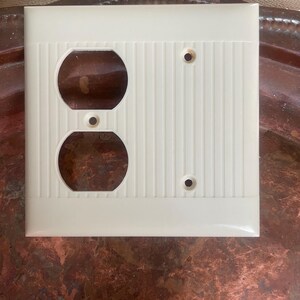 May include: Off-white electrical outlet cover with two circular openings and two screw holes. The cover has a vertical ribbed design on the right side and a smooth, glossy finish. The cover is square and appears to be made of plastic.